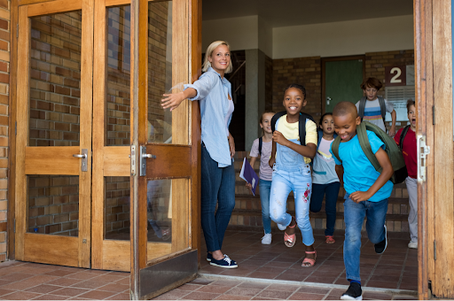 A teacher holds a wood door open as young students exist a school. They are happy because it's summer break. Everyone is smiling.
